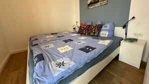 Una cama bien hecha con una colcha azul y blanca estampada, un cojín floral y una mesita de noche con una lámpara y una caja decorativa.