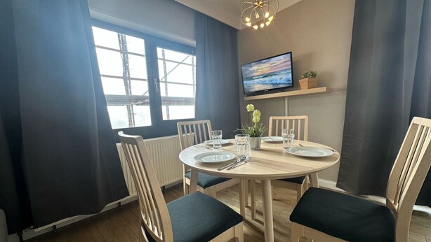 Un comedor con una mesa redonda, cuatro sillas, un televisor montado en la pared y una ventana con vistas a una obra en construcción.