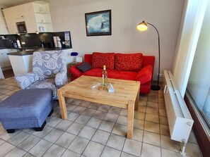 Un espacio habitable compacto con kitchenette, un sofá rojo, un sillón estampado, una mesa de centro de madera y una lámpara de pie.