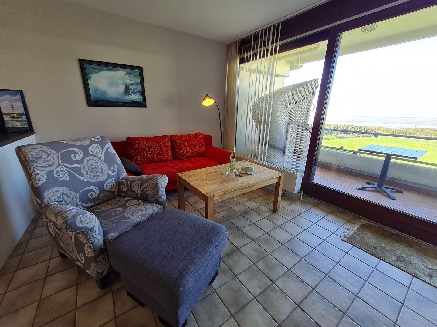 Sala de estar com sofá, poltrona e um banquinho, uma mesa de centro de madeira, e vista para o exterior através de uma porta de vidro deslizante.