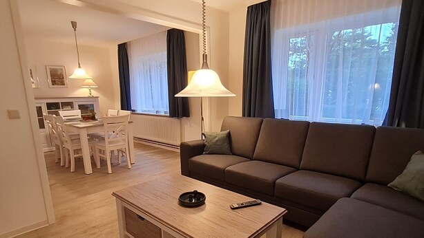 A modern living room with a brown sofa, a wooden coffee table, and a dining area with white chairs and a table. - Am Museum - Holiday cottage Am Museum (Wyk auf Föhr)