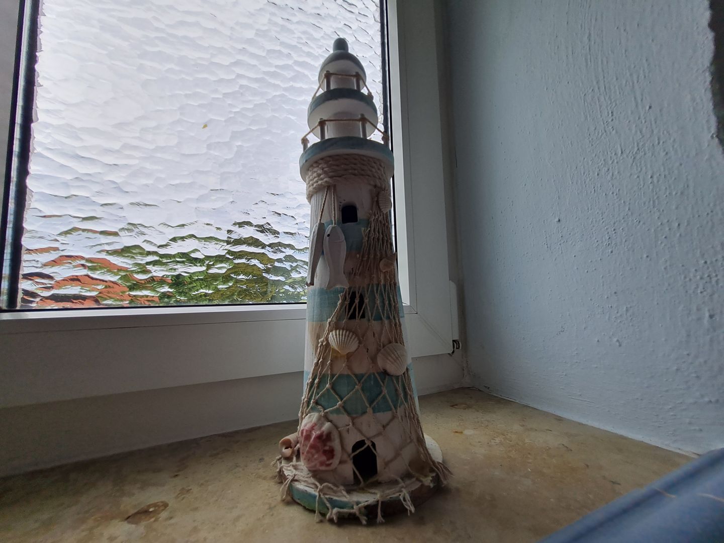 A small, decorative lighthouse model with a rope-woven base, positioned near a window overlooking a body of water.