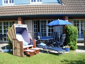 Um pátio com mobiliário de exterior, incluindo uma cadeira de vime, espreguiçadeiras e uma mesa com um guarda-sol azul.