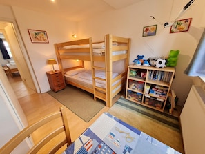 Quarto de criança com beliches, uma escrivaninha e uma estante cheia de brinquedos e livros.