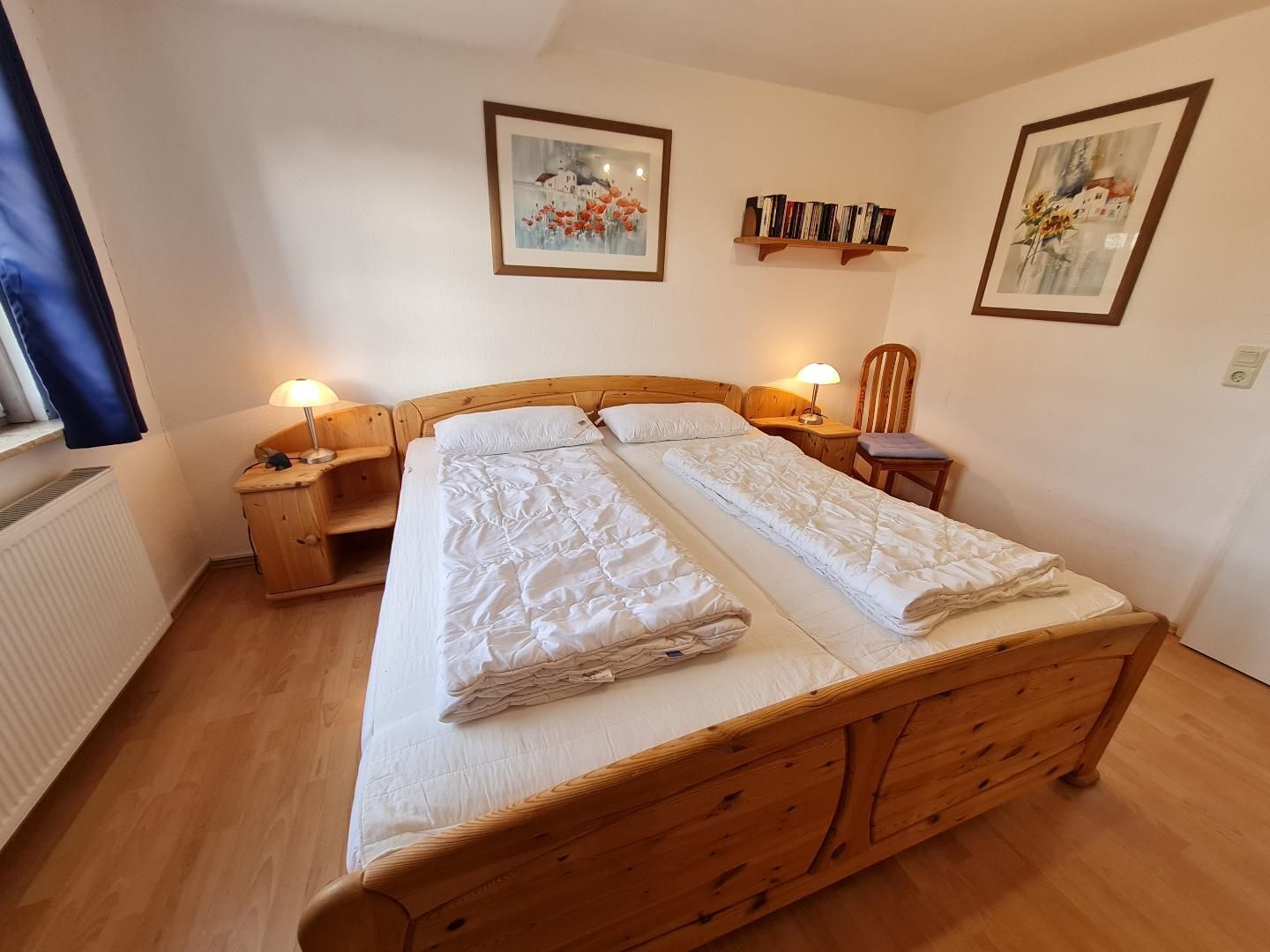 Una cama doble con cabecera de madera, dos mesitas de noche con lámparas, una silla y dos cuadros enmarcados en la pared.