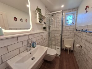 Uma casa de banho com lavatório branco, sanita e cabine de duche em vidro. Há objetos decorativos em prateleiras e uma pequena janela sobre a sanita.