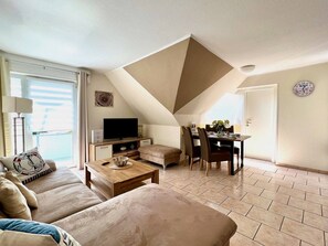 A modern living room with a sofa, coffee table, and a TV. There's a dining area with chairs and a table. The room has a sloped ceiling and a large window.