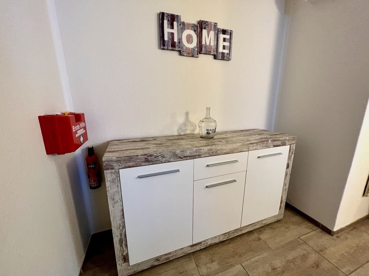 Canto de um quarto com um armário branco, uma placa de madeira acima com a palavra 'HOME' e uma caixa de alarme de incêndio vermelha na parede.