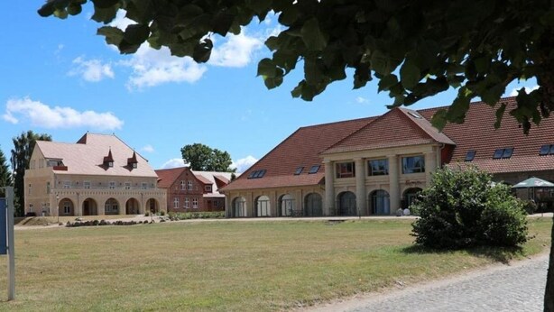 A courtyard with a cobblestone path, a grassy area, and a building with a red-tiled roof and arched windows. - Die Remise Lexington RE-03 Holiday apartment with terrace on Usedom - Die Remise (Stolpe auf Usedom)