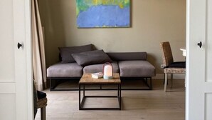 A modern living room with a grey sofa, a small wooden coffee table with a book and a lamp, a wicker chair, and a painting on the wall. - Die Remise Lexington RE-03 Holiday apartment with terrace on Usedom - Die Remise (Stolpe auf Usedom)