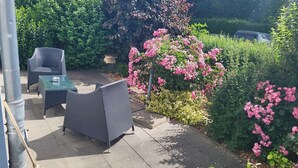 An outdoor patio area with wicker furniture and a glass table, surrounded by lush greenery and flowering plants.