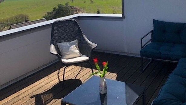 Un balcon donnant sur un champ verdoyant, une chaise, une table avec un vase de fleurs et un canapé bleu.