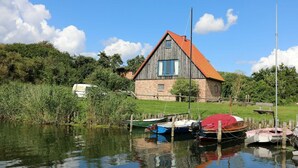 A rustic wooden house with a red roof, surrounded by a calm water body with boats, and lush greenery in the background. - Cozy vacation apartment Landhof Schwalbennest LH-204 quiet village location on Usedom (Stolpe auf Usedom)