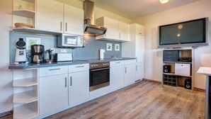 Een moderne keuken met witte kasten, een inbouwoven, een magnetron en een koffiezetapparaat. Er is een flatscreen televisie aan de muur bevestigd en een houten vloer.