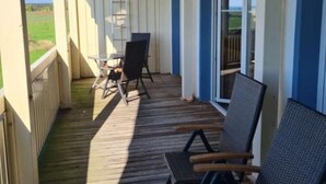 A covered outdoor deck with wooden flooring, two black chairs, a table, and a sliding glass door leading to a grassy area. - Cozy vacation apartment Landhof Weide LH-202 in a quiet village idyll on Usedom - (Stolpe auf Usedom)
