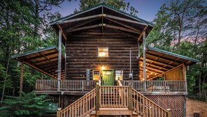 A rustic log cabin with a wooden deck and a green door, surrounded by dense forest. - 2 King Beds - Hot Tub - 10 min to parkway (Sevierville)