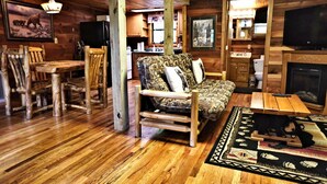 A rustic cabin interior with a wooden dining table, a patterned sofa, a fireplace, and a television. - 2 King Beds - Hot Tub - 10 min to parkway (Sevierville)
