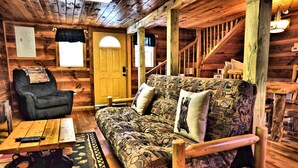A cozy cabin interior with a wooden floor, a patterned sofa, a black armchair, a wooden table, and a staircase. - 2 King Beds - Hot Tub - 10 min to parkway (Sevierville)