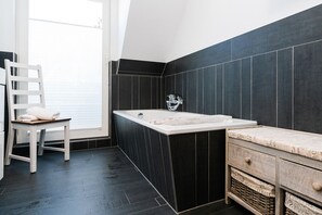 A modern bathroom with a bathtub, a white chair with towels, and dark tiled walls. - Erika - House Erika, Germany (Sylt)