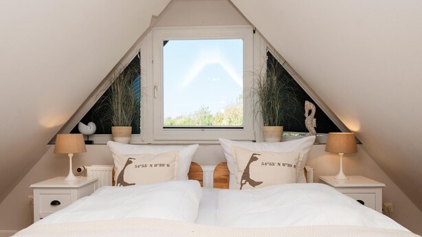 A bedroom with a sloped ceiling, a large window, two bedside tables with lamps, and two decorative pillows with coordinates. - Erika - House Erika, Germany (Sylt)