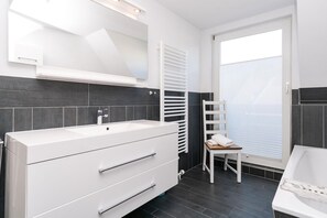 Un baño moderno con un lavamanos grande de color blanco, paredes de azulejos oscuros, una silla blanca y una ventana con persianas.