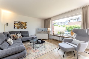 Ett modernt vardagsrum med en grå soffa, en grå fåtölj, ett soffbord och en tv på ett vitt skåp. Det finns ett stort fönster med utsikt över byggnader och grönska utanför.