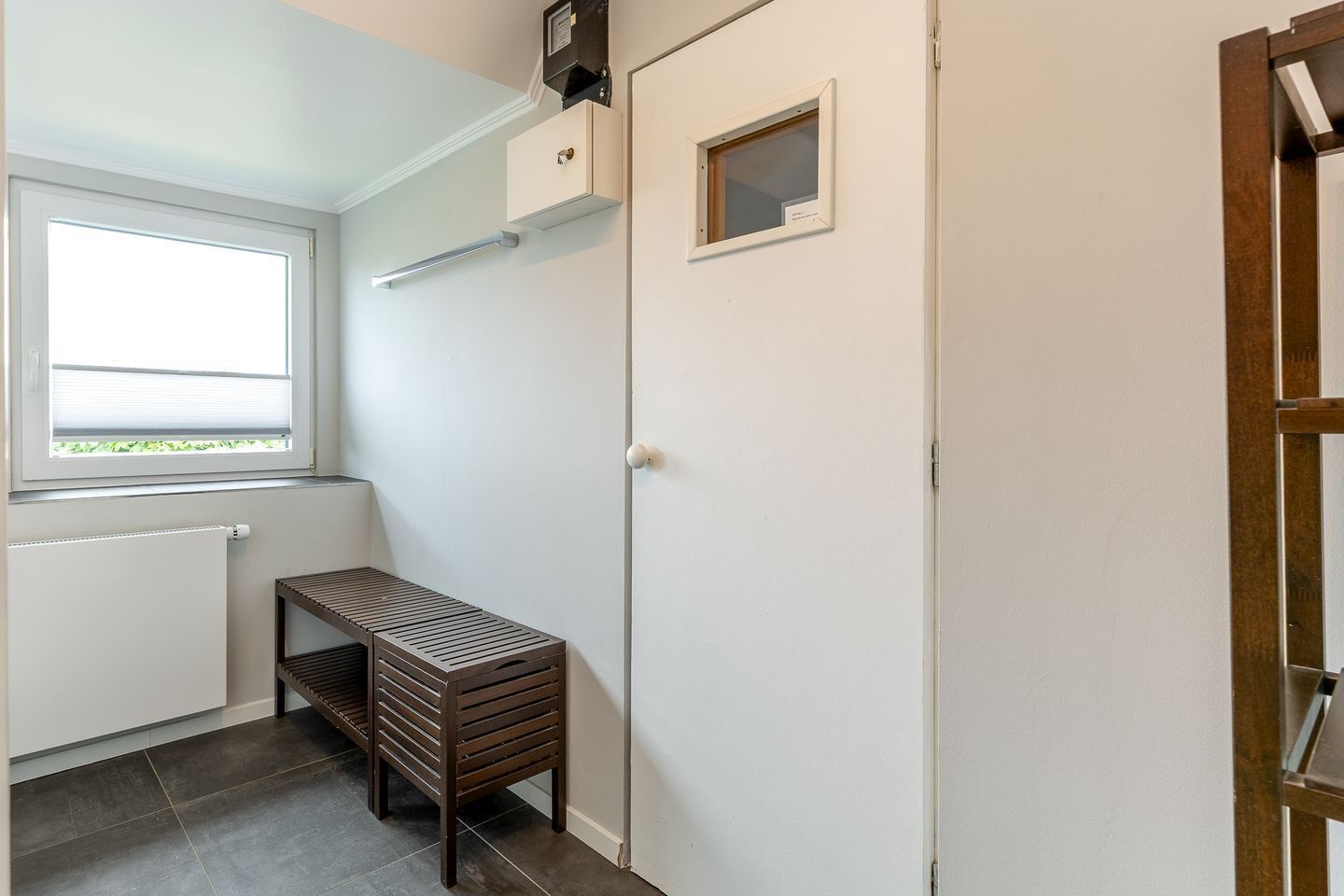 Una habitación pequeña y limpia con ventana, un radiador, un banco de madera y una puerta.