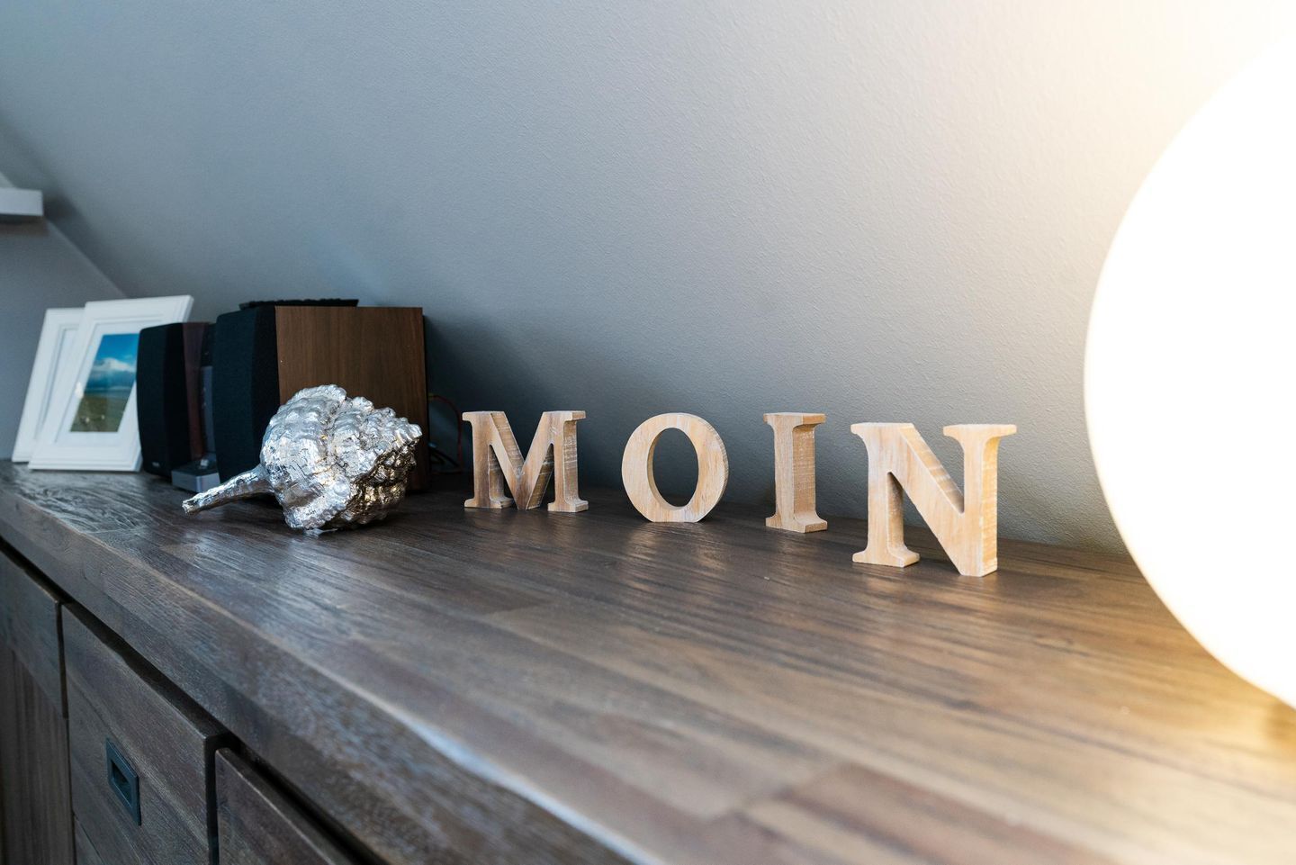 Holzbuchstaben, die "MOIN" buchstabieren, auf einem dunklen Holzbrett, mit einer dekorativen Muschel und gerahmten Bildern im Hintergrund.