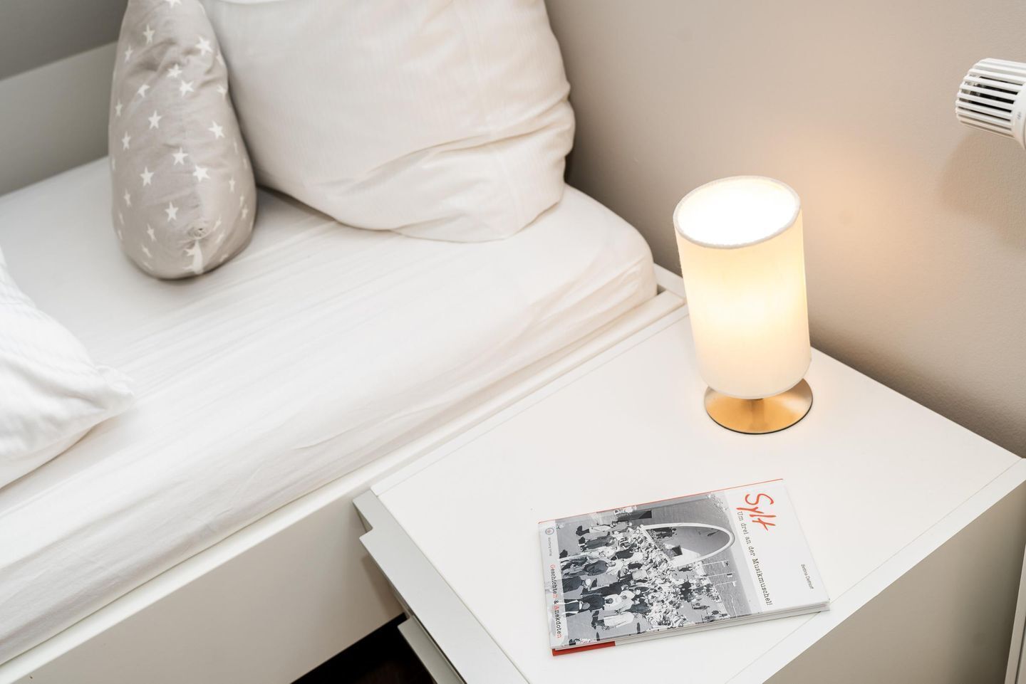 Ein ordentlich bezogenes Bett mit weißer Bettwäsche und einem Kissen mit Sternenmuster, ein Nachttisch mit einer Zeitschrift und eine beleuchtete Tischlampe.