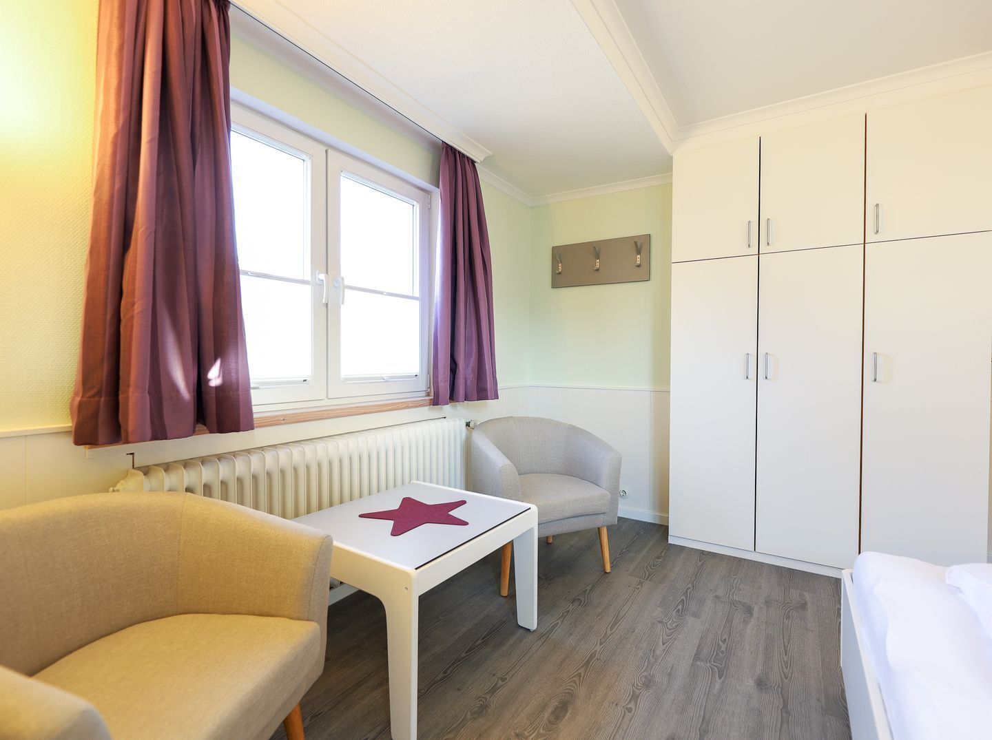 A room with a bed, a chair, a table with a star decoration, a wardrobe, and a window with curtains.