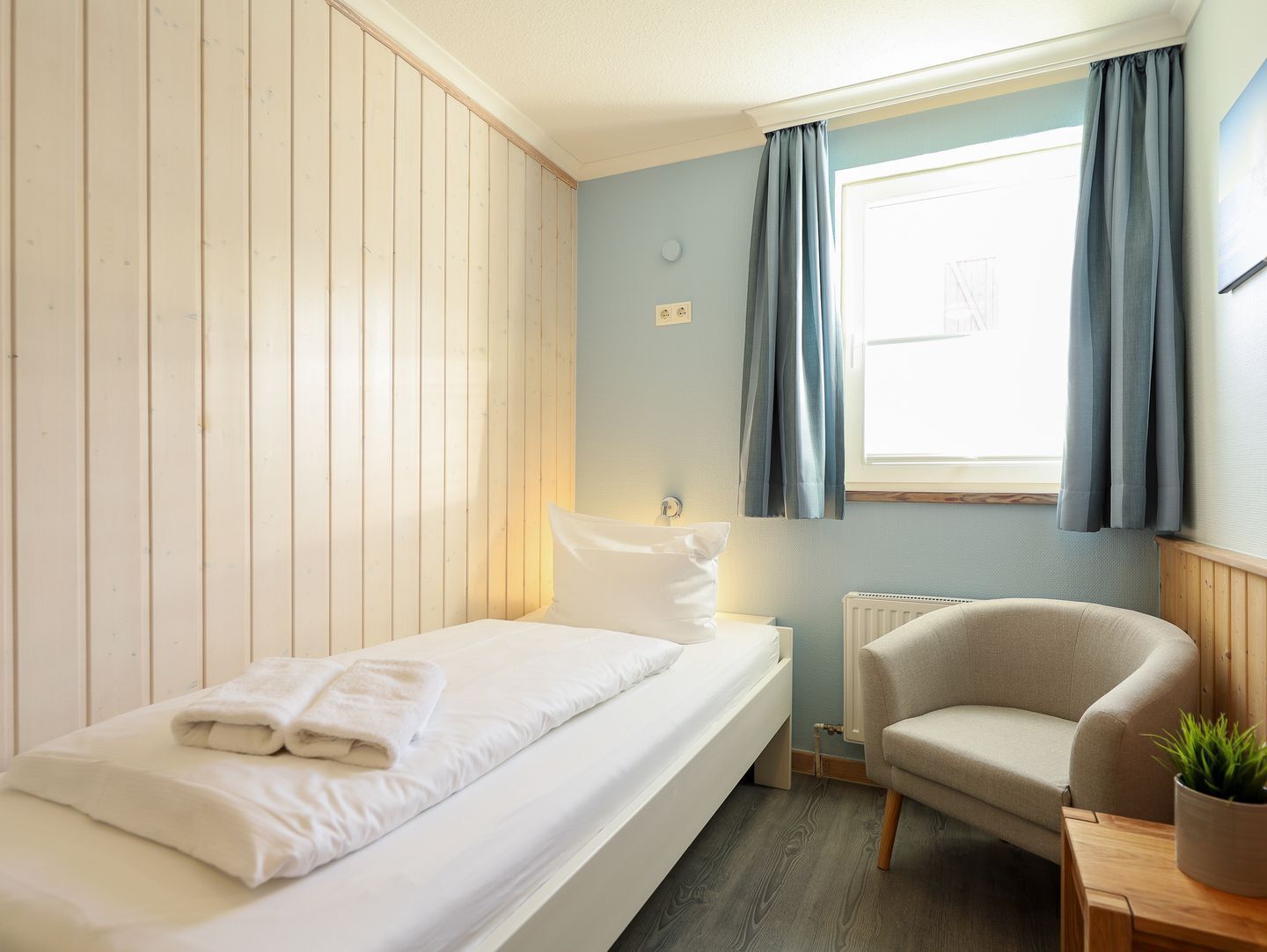 Un dormitorio pequeño y ordenado con una cama individual, una silla y un ventanal con cortinas.