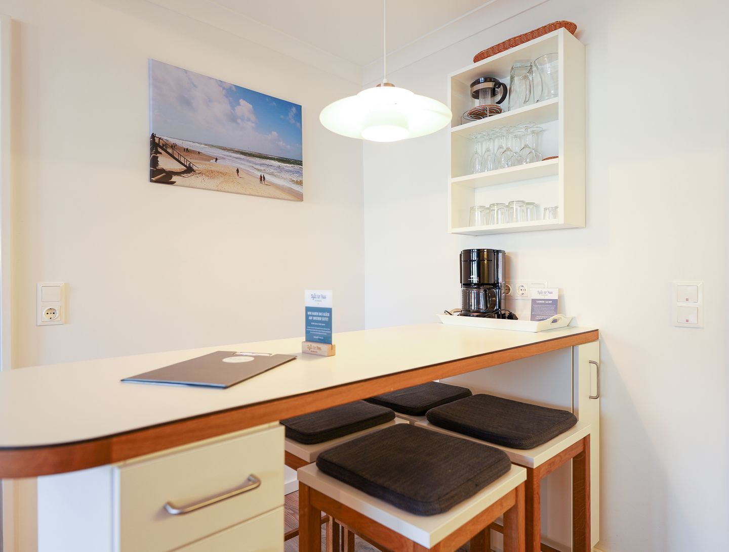 A small kitchenette with a coffee maker, a shelf with glasses, a bench, a desk with a laptop, and a beach painting on the wall.