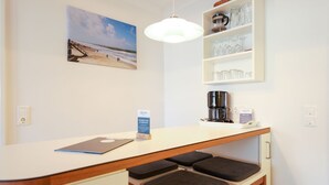 A small kitchenette with a coffee maker, a shelf with glasses, a bench, a desk with a laptop, and a beach painting on the wall. - Left - House Rolf - Left (Sylt)