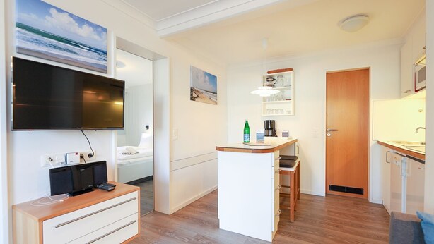 A compact hotel room with a bed, a desk, a kitchenette, and a TV mounted on the wall. - Left - House Rolf - Left (Sylt)