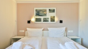 Un lit double avec du linge de lit et des serviettes blanches, deux tables de chevet, une lampe fixée au mur et un tableau représentant des paysages.