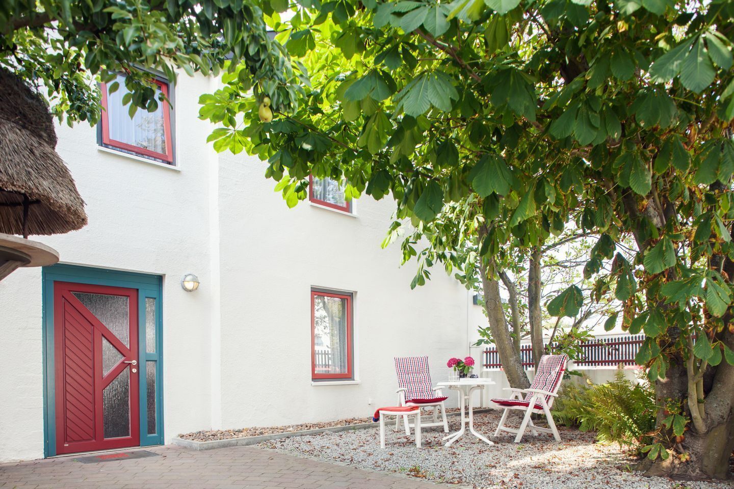 Un bâtiment blanc avec une porte et des fenêtres rouges, une toiture en chaume et des sièges extérieurs dotés de coussins à rayures rouges et blanches.