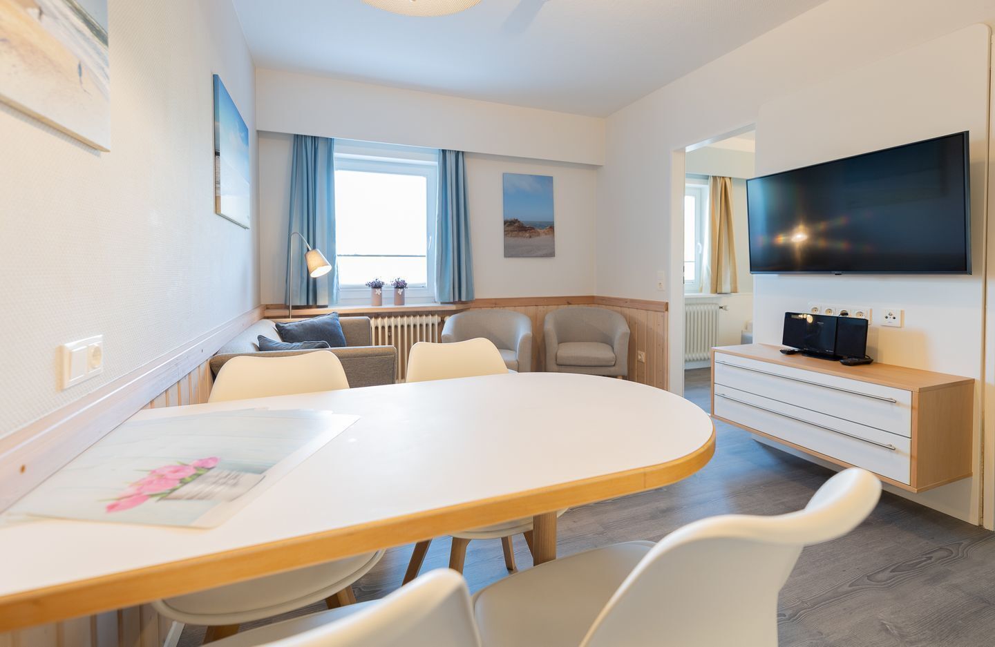 A modern hotel room with a round dining table, two armchairs, a sofa, a TV, and a small cabinet.
