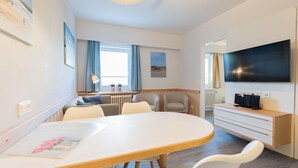 A modern hotel room with a round dining table, two armchairs, a sofa, a TV, and a small cabinet.