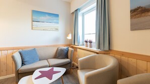 A cozy room with a sofa, armchair, and a small table with a star decoration. A window with curtains and a painting of a beach are visible.