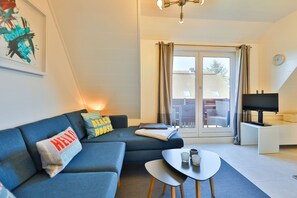 A modern living room with a blue sofa, a small table with glasses, a TV on a stand, and a sliding glass door leading to an outdoor area.