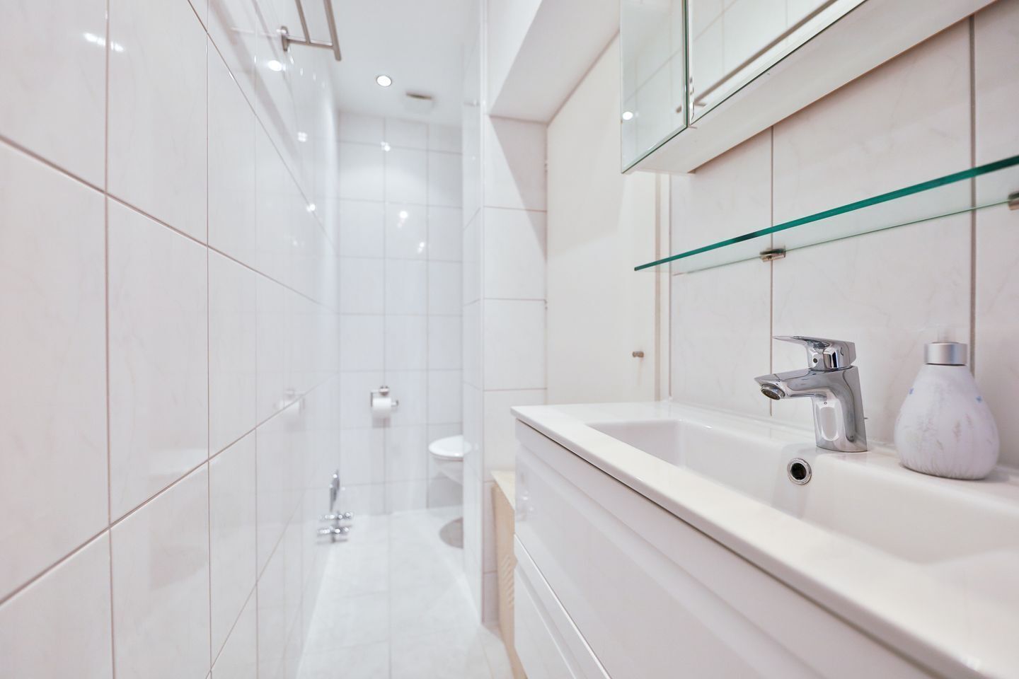 A modern bathroom with white tiles, a glass shower enclosure, a white sink with a chrome faucet, and a marble soap dispenser.
