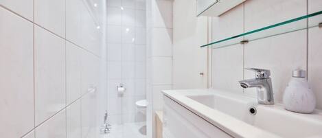 A modern bathroom with white tiles, a glass shower enclosure, a white sink with a chrome faucet, and a marble soap dispenser.