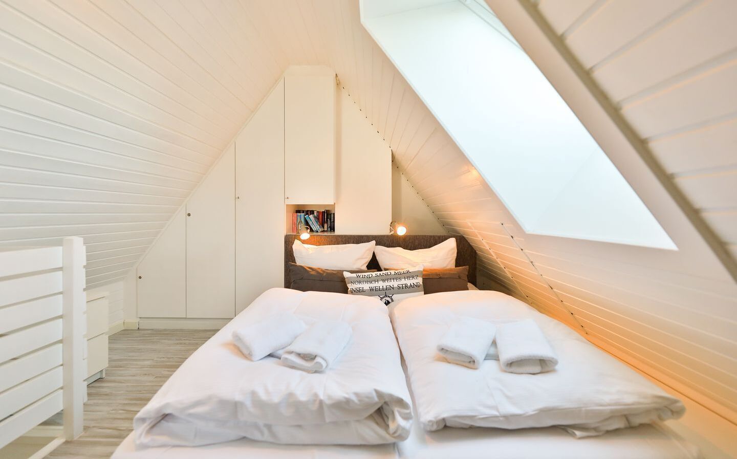 A bedroom with a sloped ceiling and a skylight, featuring a bed with white linens and pillows, a bedside table with books, and a wall-mounted shelf.