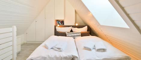 Un dormitorio con techo inclinado y tragaluz, que incluye una cama con ropa de cama y almohadas blancas, una mesita de noche con libros y un estante montado en la pared.