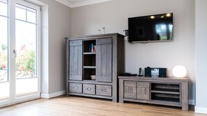 A room with a wooden cabinet, a TV mounted on the wall, and a small table with electronic devices. - Lille Kamp 1, Germany (Sylt-Ost)