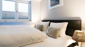 A bedroom with a large bed, a nightstand with a lamp, and a window with blinds. - Lille Kamp 1, Germany (Sylt-Ost)