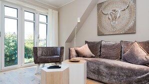 Un salon moderne avec un canapé gris, un fauteuil marron, une petite table d’appoint blanche et une grande fenêtre donnant sur un paysage verdoyant.