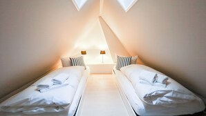 Una habitación pequeña y bien iluminada con dos camas individuales, cada una con una almohada y una toalla doblada. Hay dos lámparas de noche en una mesita entre las camas.