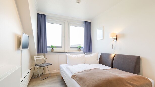 A hotel room with a bed, a chair, a desk, a TV, and a window with curtains. - Jasper - Hedighuis - Jasper in the Hedighuis (Sylt)