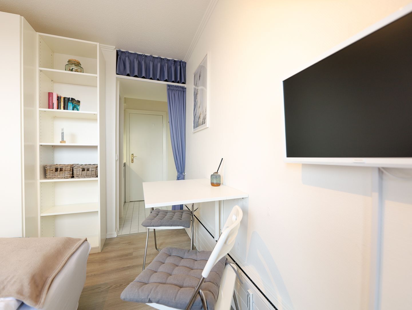 A small, well-lit room with a white desk, two chairs, a bookshelf, and a bed.